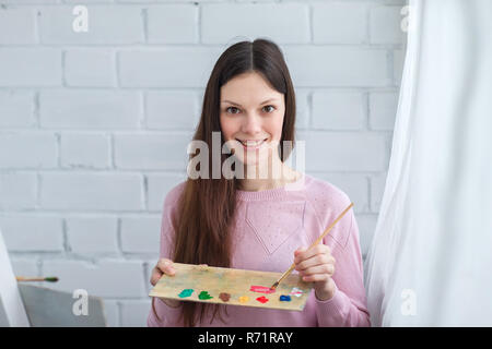Schöne junge Frau malen ein Bild auf einer Staffelei. In der Hand einen Pinsel und Palette von Farben. Stockfoto