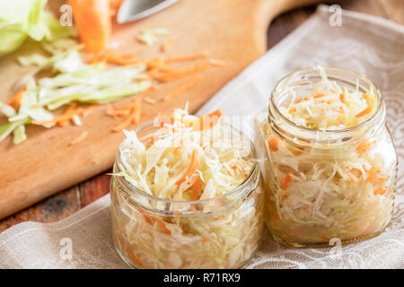 Sauerkraut in Gläsern auf dem Handtuch und Schneiden von frischem Kohl und Möhren auf Holzbrett. Stockfoto