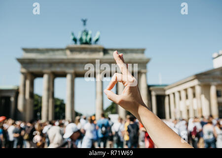 Eine Person zeigt ein Zeichen mit seinen Fingern, dass bedeutet, alles vor dem Hintergrund des Brandenburger Tor und blurry unkenntlich Menschen in Berlin in Deutschland gut ist. Stockfoto