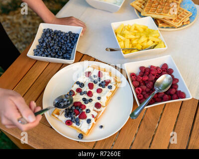 Zubereitung von hausgemachten Waffeln im Waffeleisen draußen im Garten - Frau schmückt Waffeln mit Sahne und Früchten Stockfoto