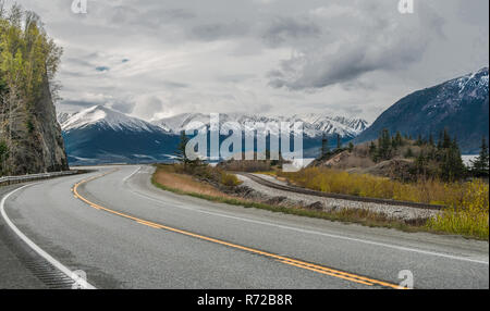 Alaska malerische Straße: Der Seward Highway Kurven unter bewölktem Himmel wie es geht durch Schnee - Berge am Rande eines Ozeans Inlet südlich von Anchorage abgedeckt. Stockfoto