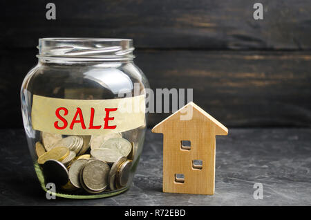 Glas Glas mit Münzen und der Inschrift "Verkauf" und einem kleinen Holzhaus. Immobilien Konzept. Verkauf von einem Haus, einer Wohnung Immobilie verkaufen. Erschwingliche Hou Stockfoto