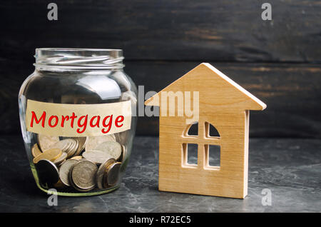 Glas Glas mit Münzen und die Inschrift 'durch Hypotheken" und einem kleinen Holzhaus. Kredit für Haus/Wohnung. Unternehmen Kredite für Immobilien Konzept. Stockfoto