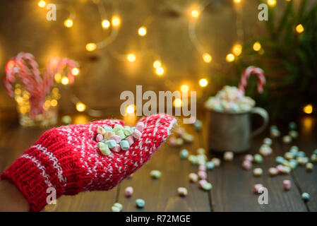 Frau Hand in Rot gestrickte Handschuhe hollding Marshmallows. Lichter in Bokeh, Becher mit Marshmallow auf Hintergrund. Weihnachten Konzept, Karte Stockfoto