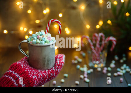 Frau Hand in Rot gestrickte Handschuhe holding Reisebecher mit Marshmallows. Lichter in Bokeh, Becher mit Marshmallow auf Hintergrund. Weihnachten Konzept, Karte Stockfoto