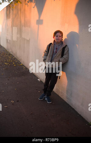 Ein elfjähriger Junge mit Rucksack lehnt sich gegen eine Wand, mit Schatten bedeckt, und wartet auf seine Schule in den frühen Morgenstunden in Montpellier zu öffnen. Stockfoto