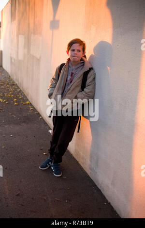 Eine zweifelhafte elf Jahre alten Jungen mit Rucksack lehnt sich gegen eine Wand, mit Schatten bedeckt, und wartet auf seine Schule in den frühen Morgenstunden zu öffnen, Frankreich Stockfoto