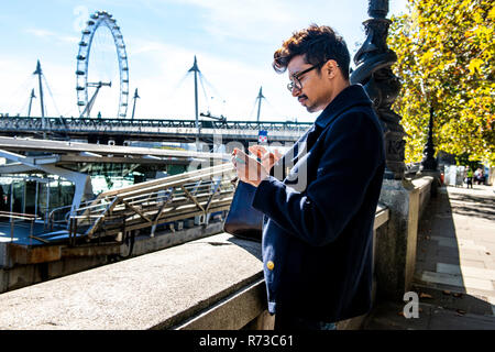 Geschäftsmann mit Smartphone auf Embankment, London Eye und Hungerford Brücke im Hintergrund, London, UK Stockfoto