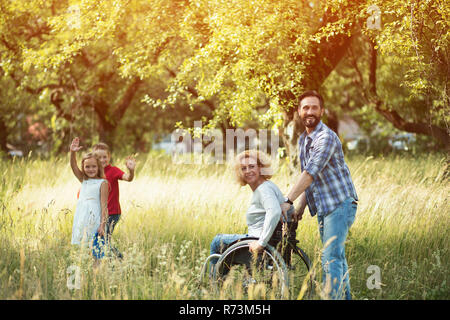 Schöne Frau im Rollstuhl mit ihrem Ehemann Die Schiebegriffe Blick zurück und lächelt Jemand. Zwei reizende Kinder sind wellenartig. Stockfoto