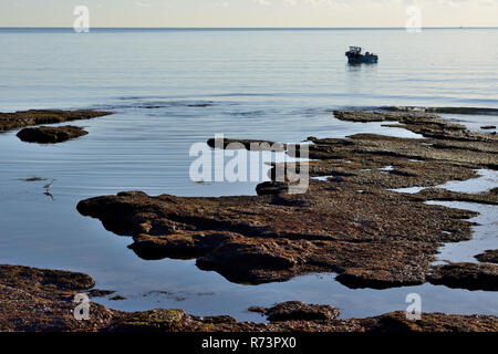 Ein kleines Fischerboot und ein kleiner Reiher Angeln unter exponierten Felsen entlang der Küste bei Ebbe. Stockfoto