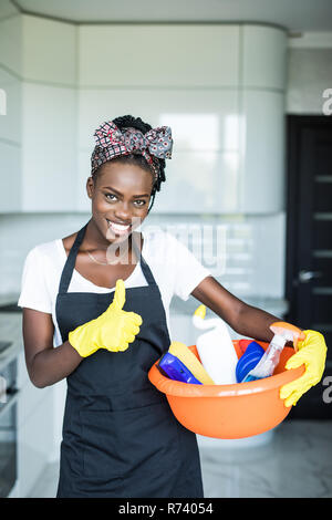 Junge afrikanische Frau mit Reinigung Korb mit gelb Handschuhe, die Daumen oben zu Hause angezeigt Stockfoto