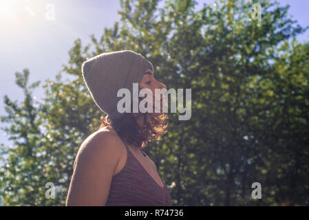 Junge Frau in Strickmütze Kontemplation vor baum laub, beleuchteter Kopf und Schulter Profil Stockfoto