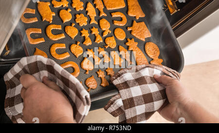Baking of Christmas gingerbreads in kitchen oven. Xmas or New Year sweets. Various shapes on hot smoking cookie sheet. Cook's hands, cloths, hot steam. Stockfoto