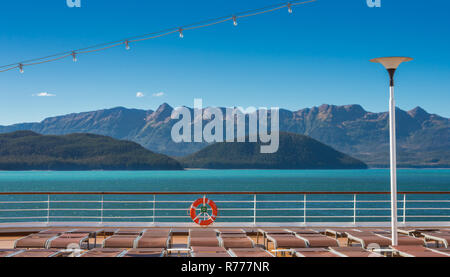 Leere Kreuzfahrtschiff Liegestühle, alle aufgrund der hohen Wind- und orange Schwimmweste am Geländer unter hellen Alaska Sonnenschein abgesenkt. Grüne Berghänge Stockfoto