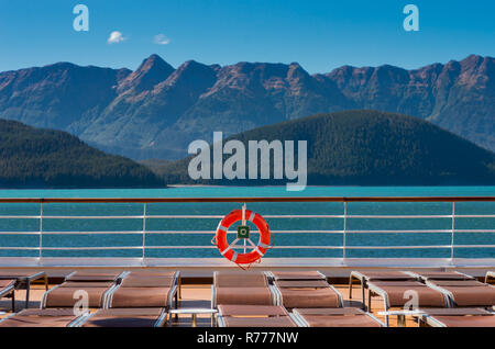 Leere Kreuzfahrtschiff Liegestühle, alle aufgrund der hohen Wind- und orange Schwimmweste am Geländer unter hellen Alaska Sonnenschein abgesenkt. Grüne Berghänge Stockfoto