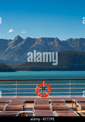 Leere Kreuzfahrtschiff Liegestühle, alle aufgrund der hohen Wind- und orange Schwimmweste am Geländer unter hellen Alaska Sonnenschein abgesenkt. Grüne Berghänge Stockfoto