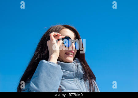 Schließen Portrait von Eine langhaarige Brünette in gespiegelten blau Sonnenbrille und Winterkleidung gegen einen strahlend blauen Himmel. Winterurlaub, Warten auf Frühling Stockfoto