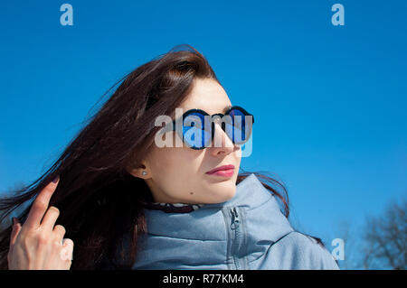 Schließen Portrait von Eine langhaarige Brünette in gespiegelten blau Sonnenbrille und Winterkleidung gegen einen strahlend blauen Himmel. Winterurlaub, Warten auf Frühling Stockfoto