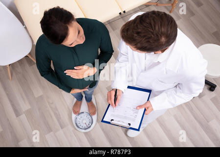 Männlicher Arzt Messen Gewicht der schwangeren Frau auf der Waage Stockfoto