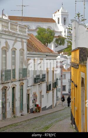 TAVIRA, PORTUGAL - 17. NOVEMBER 2018: die typischen Fassaden entlang einer steilen gepflasterten Straße mit Santa Maria Castelo Kirche im Hintergrund tun Stockfoto