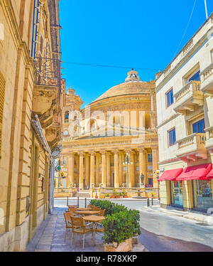 MOSTA, MALTA - 14. Juni 2018: Die schmalen schattigen Straße mit Outdoor Cafe öffnet den Blick auf die geschäftige Rotunde Platz mit seinem Wahrzeichen - Basilika der Stockfoto