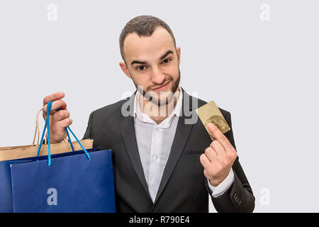 Schöne und selbstbewusste junge Mann im Anzug steht und zeigt Kreditkarte. Er sieht gerade. Junge Mann hat Einkaufstüten in der Hand. Auf weissem Hintergrund. Stockfoto