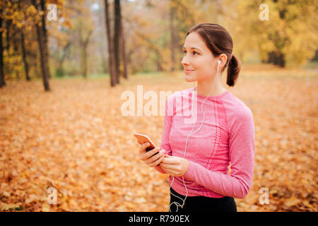 Freundliche und nette junge Frau steht allein im Park. Jogger Musik hören über Kopfhörer. Sie hält das Telefon in Händen. Junge Frau lächelt. Stockfoto
