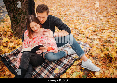 Junger Mann in schwarzer Kleidung sitzt auf Decke und Blick auf Buch Frau hält in den Händen. Sie sah es. Junge Mann Frau umarmen. Stockfoto