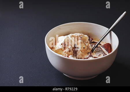 Einfache weiße Keramik Schüssel mit Vanilleeis mit Likör und zerkleinerte Schokolade Cookies auf schwarz matt lackiert Tisch gefüllt. Stockfoto