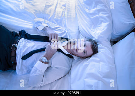 Attraktive erschöpft Geschäftsmann liegend auf Bett bis in die Kamera schauen, als er seine Krawatte unfastens Stockfoto