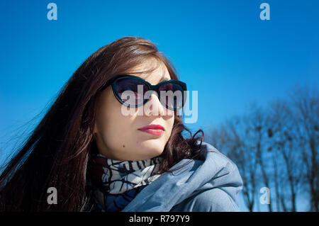 Schließen Portrait von Eine langhaarige Brünette in gespiegelten blau Sonnenbrille und Winterkleidung gegen einen strahlend blauen Himmel. Winterurlaub, Warten auf Frühling Stockfoto