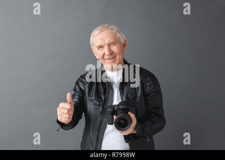 Älterer Mann in der Lederjacke stehen isoliert auf grau, die Kamera mit dem Daumen bis heiter Stockfoto
