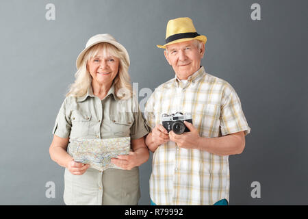 Ältere Touristen in Strand hüte Studio stehen isoliert auf Grau holding Kamera und Karte heiter Stockfoto