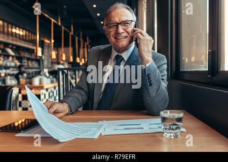 Lächelnd Reife Unternehmer sitzen im Café sprechen über Telefon mit dem Dokument in der Hand. Gerne ältere Unternehmer telefonieren zum Client. Stockfoto