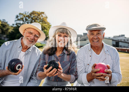 Lächelnde ältere Leute sitzen in einem Rasen, Boccia. Zwei Männer und eine Frau genießen das Spiel Boule sitzen zusammen in einem Park. Stockfoto