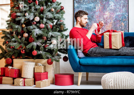 Man-Eröffnung ein Geschenk Box auf der Couch liegen während der Winterferien mit Weihnachtsbaum zu Hause Stockfoto