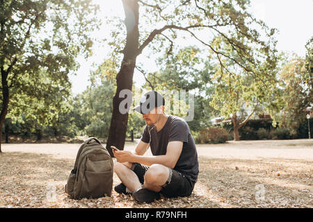 Junge positive Kerl oder Student mit dem Rucksack sitzt und das Hören von Musik oder Podcasts oder Radio in einem Stadtpark. Lebensstil Stockfoto