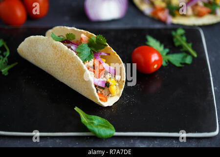 Mexikanische Tacos mit Schweinefleisch, Gemüse und Gewürze auf einem schwarzen Stein Platte auf einem dunklen Hintergrund mit Zutaten für Tacos Menü und Restaurant Konzept. Stockfoto