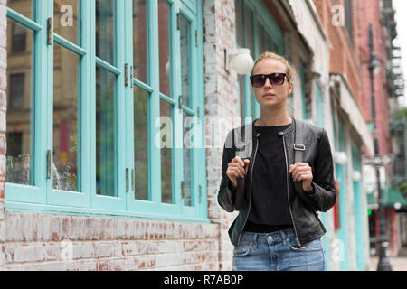 Modische junge Frau in schwarzer Lederjacke und beschäftigt mit ihrem Mobiltelefon beim Gehen eine Straße der Stadt Stockfoto