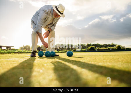 Älterer Mann in Hut Bücken eine Boule stehend auf einem Spielplatz. Älterer mann Boule spielen in einem Rasen mit Sonne in den Hintergrund. Stockfoto