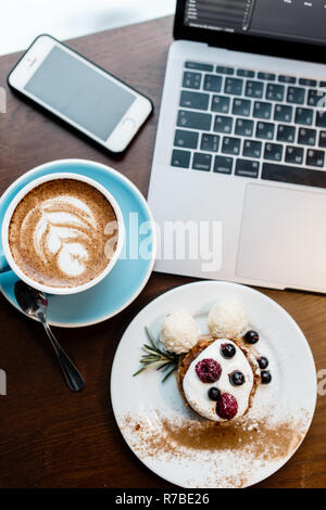 Blick von oben auf die Designer statt. Freie Arbeitsfläche. Notebook mit Handy, Tasse Kaffee und Dessert auf Holztisch. Stockfoto
