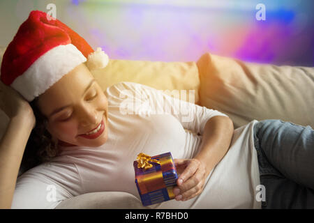 Glückliche afroamerikanische Frau tragen Santa hat an Weihnachten Geschenk im Sofa zu Hause liegen. Unscharfer Hintergrund mit Bokeh leuchtet. Stockfoto