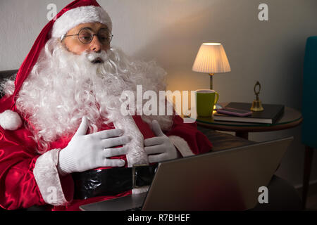 Vorderansicht von Santa Claus (oder Vater Weihnachten) sitzen im Sofa zu Hause mit Laptop für Kommunikation und Freizeit. Stockfoto