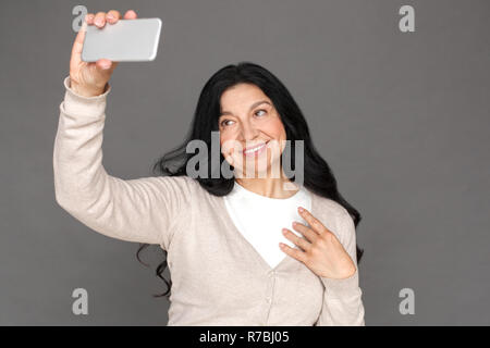 Freestyle. Reife Lady stehen isoliert auf Grau unter selfie auf dem Smartphone lächelt Fröhlich Stockfoto