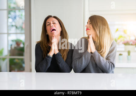 Schöne Familie von Mutter und Tochter zusammen zu Hause betteln und mit Händen gemeinsam beten mit der Hoffnung Ausdruck auf dem Gesicht sehr emotional und worri Stockfoto