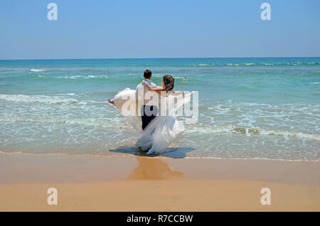 Der Bräutigam in einem Anzug trägt auf seinen Händen die Braut in einem Hochzeitskleid in den Gewässern des Indischen Ozeans. Hochzeit und Flitterwochen in den Tropen auf der i Stockfoto