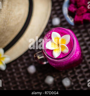 Red Dragon Obst im Glas dekoriert mit Plumeria Flower. Hut, Eiswürfel, Dragon Fruit über dunkle braune Oberfläche schneiden. Ansicht von oben. Stockfoto