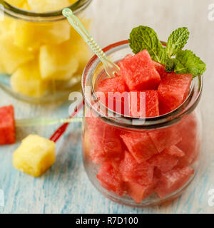 Würfel schneiden der rote und gelbe Wassermelone in Gläsern mit Minze und Kunststoff canape holt über Weiß und Blau gewaschen Holztisch. Von der Seite. Close-up. Stockfoto