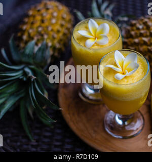 Zwei Gläser mit frischem Ananassaft mit Plumeria Blumen, zwei ganze Ananas und Baum großen Stücke auf einem runden hölzernen Fach. Stockfoto
