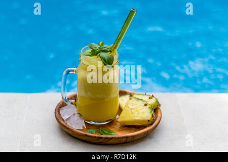 Glas frischen Ananassaft mit Glas Stroh, serviert mit zwei Stücken von Ananas, Würfel aus Eis und basilikumblätter am Rand des Pools. Stockfoto